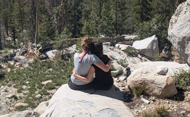 Two people are sitting on a rock, embracing each other. They appear to be in a mountainous or forest area, surrounded by trees and large rocks. The person on the left is wearing a light-colored shirt, while the person on the right is wearing a dark-colored shirt. They are sitting close together, with their arms around each other, suggesting a close relationship or moment of comfort and connection in a natural setting.